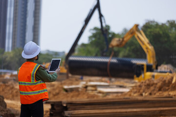 A construction engineer wearing a safety vest and hard hat monitors site progress using a tablet and phone. Heavy machinery and large drainage pipes are being installed in the background