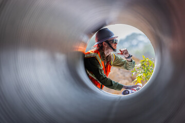 A female engineer wearing a safety vest, hard hat, and sunglasses examines a large drainage pipe...