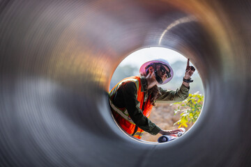 A female engineer wearing a safety vest, hard hat, and sunglasses examines a large drainage pipe from the inside perspective. She holds blueprints and inspects the structure at a construction site
