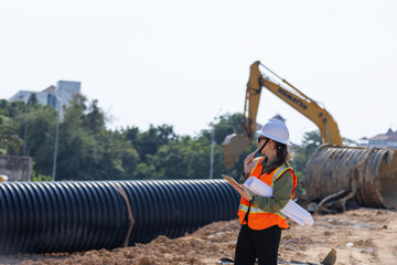 A female engineer wearing a safety vest and hard hat inspects a construction site with large...
