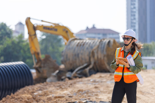 A female engineer wearing a safety vest and hard hat inspects a construction site with large drainage pipes and an excavator in the background. She holds blueprints and a tablet, documenting progress