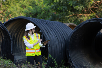 A female engineer inspects a damaged large drainage pipe outdoors. She wears a safety vest, hard...