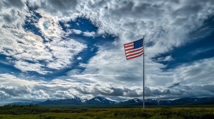 Fototapeta premium An American flag flew high against a cloudy summer sky, with Alaskan mountains in the distance. 