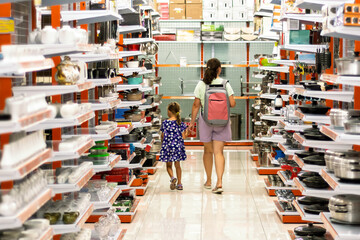 A woman and a young girl walk hand in hand through the kitchenware aisle of a bright store. The shelves are filled with cookware and home goods.