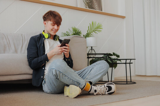 Young smiling man student with yellow headphones using phone at living room. - Powered by Adobe