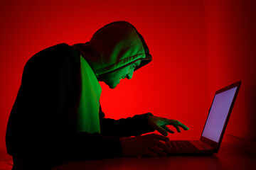 Individual Wearing Hoodie Engaged in Computer Activity With Red Background at Night