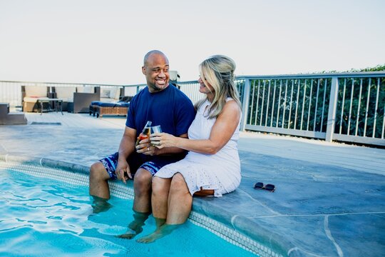 A couple enjoying a poolside moment, sitting by the water. They are smiling and holding drinks, creating a relaxed and joyful atmosphere by the pool. Couple sitting together, diverse couple.