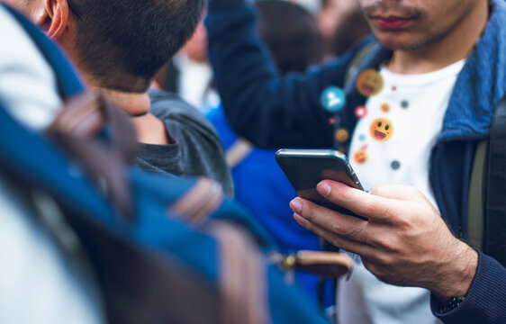 A young man interacts with his smartphone in a crowded public setting, with digital emojis symbolizing online communication and social media engagement.  
