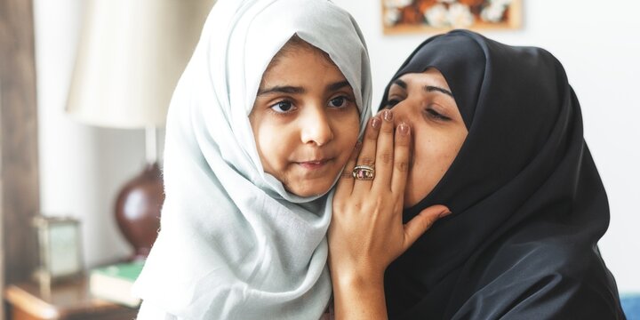 A loving Muslim mother and her young daughter share a tender moment. The mother, wearing a black hijab, whispers to her daughter in a light gray hijab, showcasing their close bond.