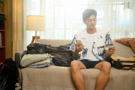 Young man in summer clothes holding smartphone and credit card sitting on couch with packed luggage