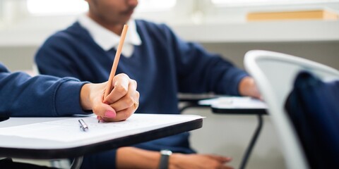 Students in a classroom taking notes. Focus on a hand writing with a pencil. Classroom setting with desks and students engaged in learning activities. Male students in classroom, education concept.
