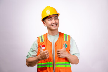 An Asian male construction worker wearing an orange safety vest and a yellow hard hat gives a thumbs-up with a confident smile. The background is white