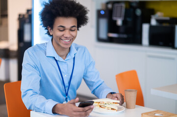 Fototapeta premium Man feeling hungry enjoying snack while browsing internet on his smartphone in office kitchen