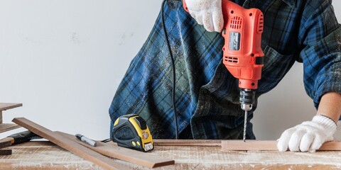 Woman using a red drill on wood, wearing gloves and a plaid shirt. Woodworking tools like a tape measure and pencil are visible. DIY woodworking scene. Woodworking and construction work concept.