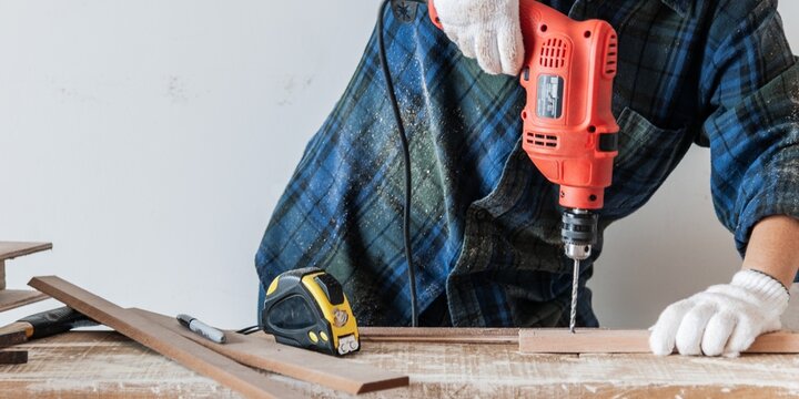 Woman using a red drill on wood, wearing gloves and a plaid shirt. Woodworking tools like a tape measure and pencil are visible. DIY woodworking scene. Woodworking and construction work concept.