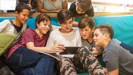Group of teenage boys sitting on a couch, looking at a tablet together, smiling and engaged. Casual setting with a focus on friendship and technology. Teenage boys on social media on a tablet.
