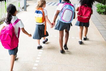 Obraz premium Four young girls with backpacks walk together on a path. School uniforms, backpacks, and friendship are highlighted in this cheerful scene. Cute little kids with backpacks in school.