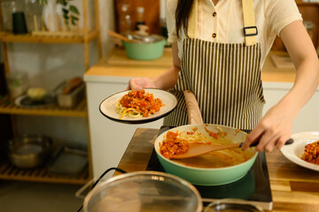 Woman serving freshly cooked spaghetti with tomato sauce from pan onto plate in a home kitchen