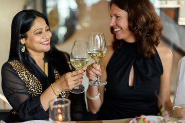 Two women enjoying a toast with wine glasses, smiling and celebrating. They are seated together, sharing a joyful moment with wine and laughter. Friends at festive dinner diverse women with wine.