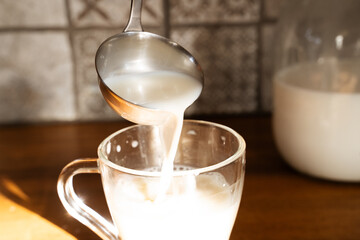 Pour fresh homemade milk into a glass with a ladle. A large glass jar filled with milk sits on a wooden surface in the background. Rustic, traditional, natural living, dairy products, milk day.