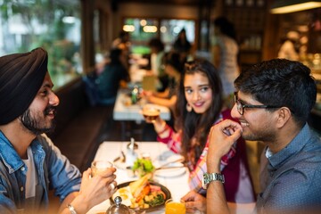 Group of friends enjoying a meal at a restaurant. Lively conversation, sharing food. Warm atmosphere, friends dining, enjoying time together. Young Indian friends eating lunch at restaurant.