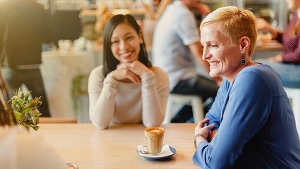 Two women, one Asian and one Caucasian, enjoy coffee together in a cozy cafe. The Asian woman...