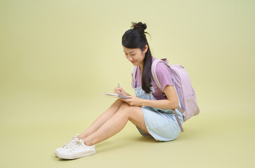 Obraz premium Young girl with backpack happily writing notes in a notebook on a pastel green background