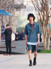 Obraz premium Portrait of handsome Chinese young man wearing short sleeved shirt and shorts posing in the street, young guy with black curly hair with urban background. male fashion, cool Asian young man lifestyle