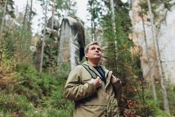 Fototapeta premium A tourist with a backpack stands against the background of a mountains and forest. Young 30s blonde man hiking in nature. Travel, adventure. Concept of an active lifestyle. High quality photo.