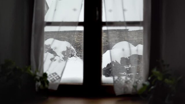 Rebord de fen&ecirc;tre avec plantes et rideaux vue sur un jardin de montagne sous la neige avec vieille grange en pierre et tas de bois