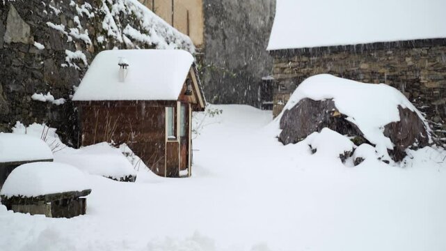 Petit chalet en bois de jardin sous la neige dans une court int&eacute;rieur en montagne avec des tas de bois et vieille grange en pierre et de la neige qui tombe