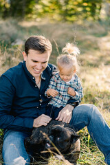 Fototapeta premium Little girl sits on the lap of her dad stroking the dog on the lawn