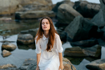Woman in white dress sitting on seaside rock, gazing out at ocean waves crashing on rocks