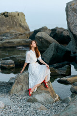 Serene woman in white dress and red shoes sitting on coastal rock overlooking ocean view