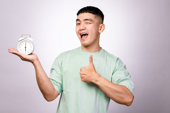 A cheerful Asian man in a mint green shirt holds a white alarm clock and gives a thumbs-up, winking at the camera. Captured against a clean white background, symbolizing good time management