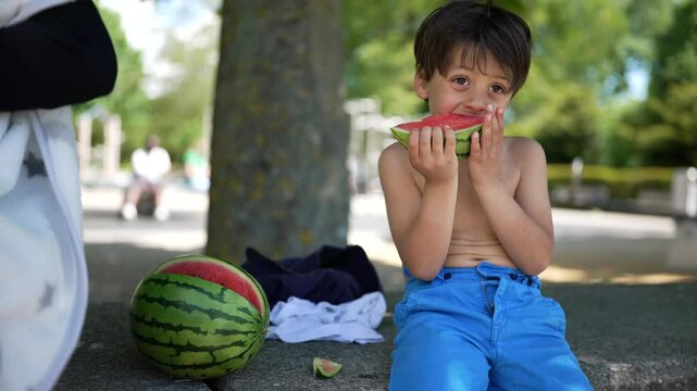Boy enjoying a slice of watermelon, sitting shirtless in the park, surrounded by nature, wearing blue pants, savoring a juicy and healthy summer snack, leisure time on a sunny day