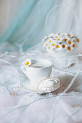 A white cup with a daisy on its saucer sits gracefully on a light fabric background. Surrounding it are subtle hues and a floral arrangement, creating a peaceful ambiance