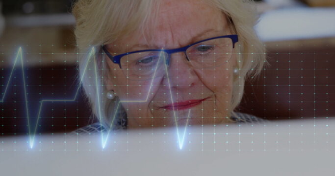 Image of cardiograph over senior caucasian woman using laptop