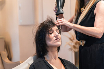 Naklejka premium Young pretty woman is sitting in the armchair in a beauty salon getting her hair dried by a smiling professional stylist