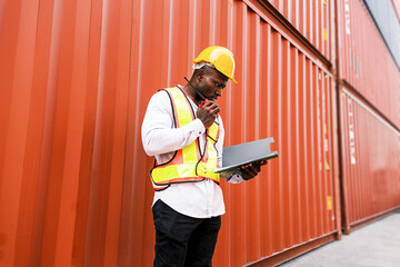 An African logistics worker wearing a hard hat and safety vest inspects shipping containers while using a radio and clipboard. Freight management, transportation, and global trade operations in action