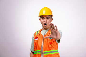 An Asian male construction worker wearing an orange safety vest and a yellow hard hat extends his hand forward in a stopping gesture with a serious expression. The background is white