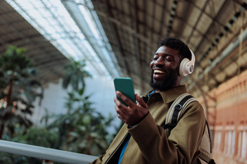 Tourist wearing headphones is using smart phone and smiling in airport or train station