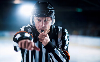 A focused referee in a striped uniform blowing a whistle and pointing, making an official call during a dynamic ice hockey game on the ice rink
