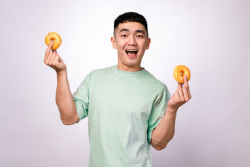An Asian man in a mint green shirt is holding two glazed donuts on his fingertips while smiling widely. He is looking happy and playful against a plain white background.