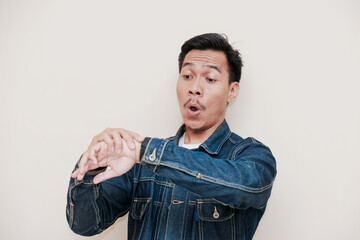 Obraz premium A Southeast Asian man wearing a denim jacket shocked expression to see watch on hand against a white background. This image is perfect for content related to time management