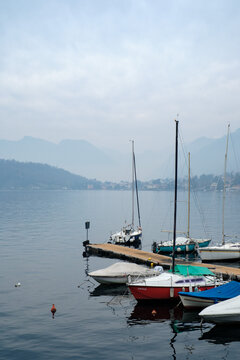 Tranquil lakeside marina with moored sailboats, calm water reflections and misty mountain backdrop creating a peaceful nautical atmosphere