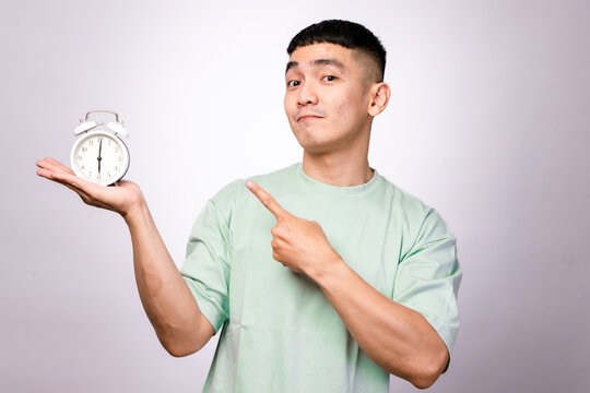 A smiling Asian man in a mint green shirt holds a white alarm clock and points at it with a cheerful expression against a white background, symbolizing time management, punctuality