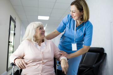 A nurse smiles warmly while assisting an elderly woman in a wheelchair at a medical clinic, creating a friendly environment