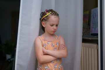 A small, cute, sad girl with colorful tiny braids stands against the backdrop of a white curtain at...