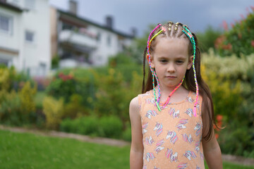 A sad, lonely, little cute girl in a dress with colorful small braids and a missing front tooth in...
